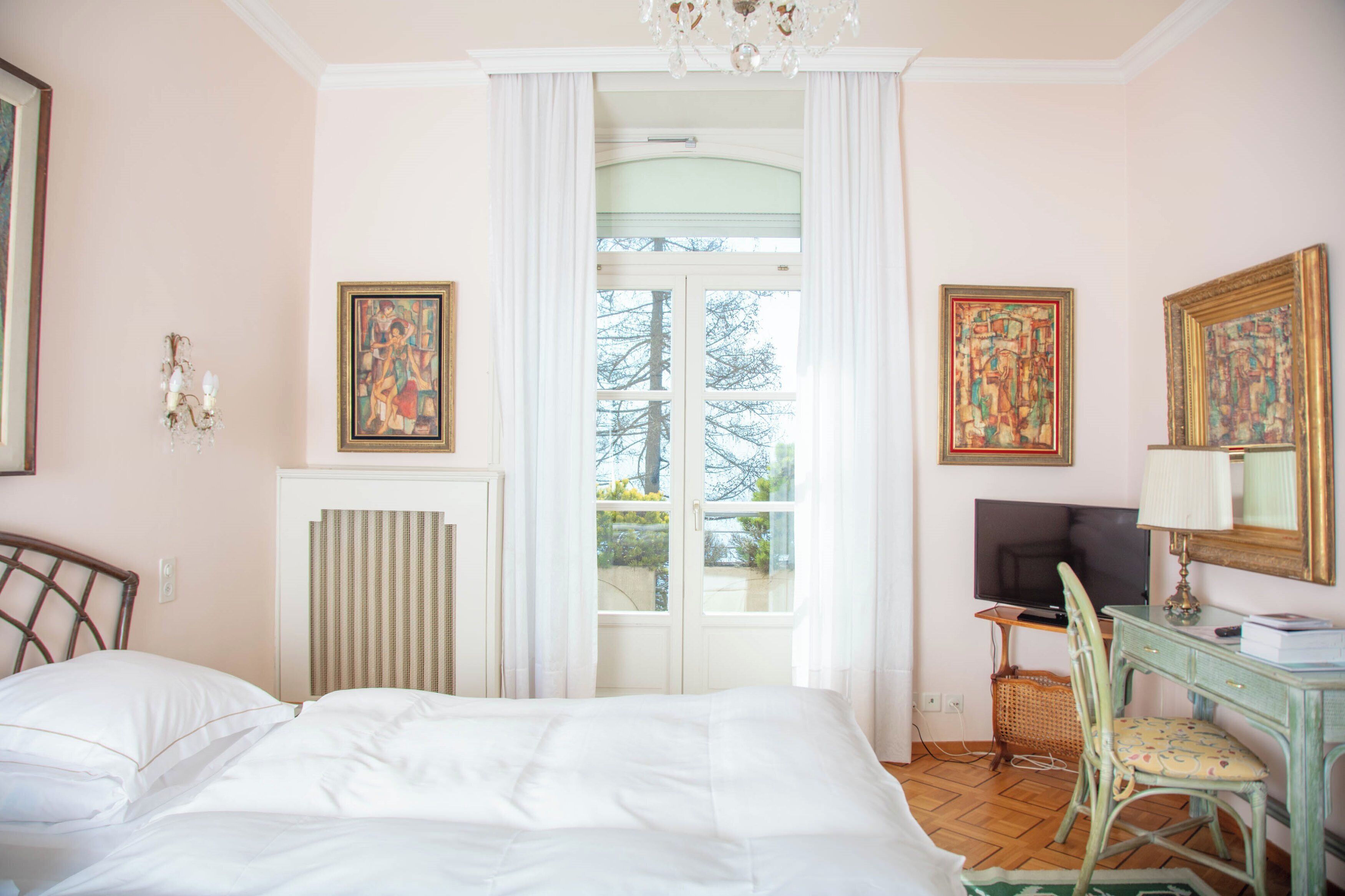 A bedroom with a bed, a desk with a chair, a television, and two paintings on the wall.