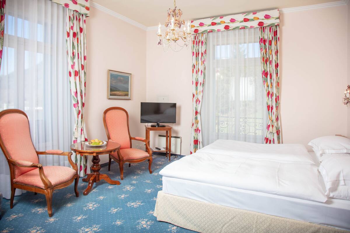 A hotel room with a bed, two chairs, a small table with a bowl of fruit, a television, and a chandelier.