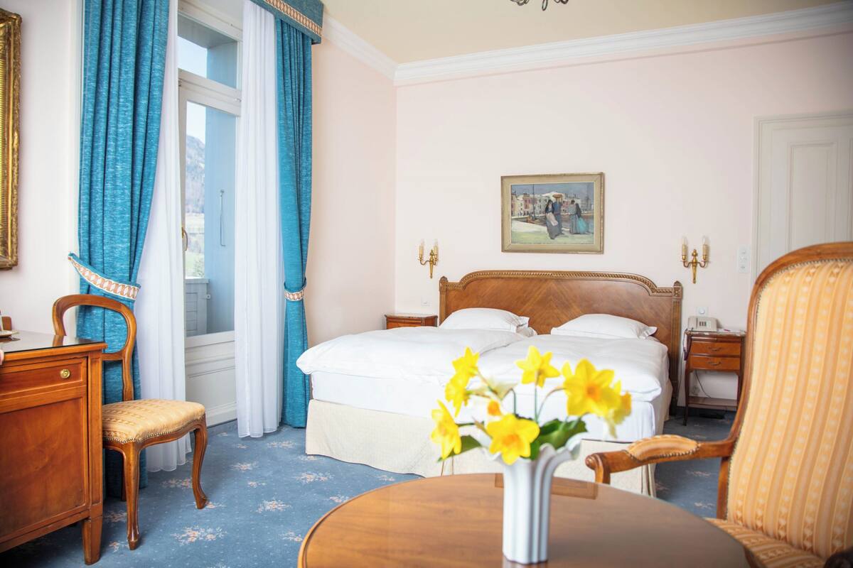 A hotel room with a large bed, a wooden desk, a chair, a small table with a vase of flowers, and a painting on the wall.