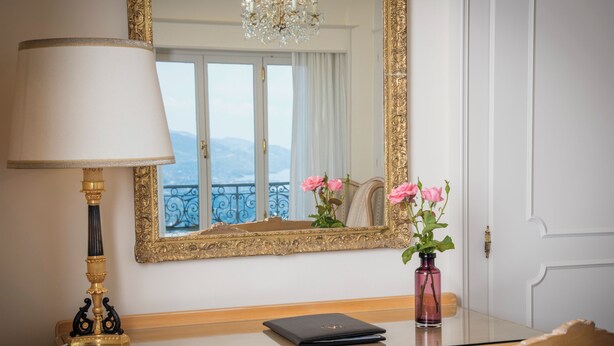 Habitación de hotel con escritorio de madera, computadora portátil negra, florero rosa con flores, espejo con marco dorado y candelabro.