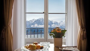 A dining table with a fruit bowl and a potted plant, a window with a view of snow-capped mountains and a body of water, and curtains on either side. - Hotel Victoria (Montreux)