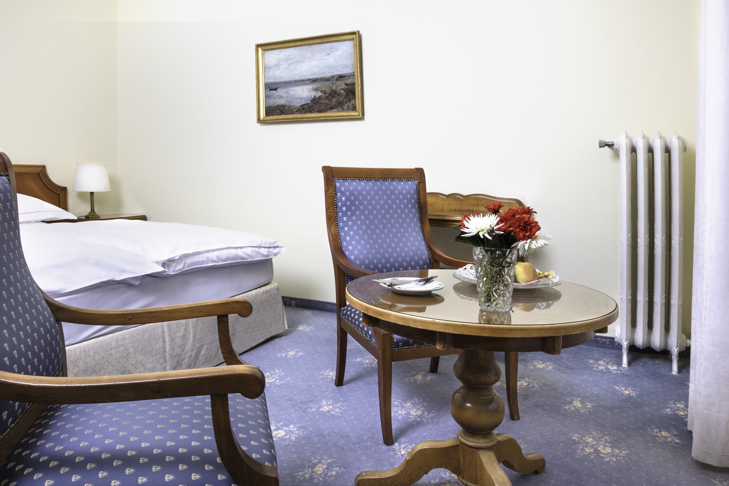 Una habitación de hotel con cama, dos sillones, una mesa de comedor con un jarrón de flores, un cuadro en la pared y un radiador.