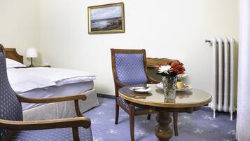 A hotel room with a bed, two chairs, a dining table with a vase of flowers, a painting on the wall, and a radiator.