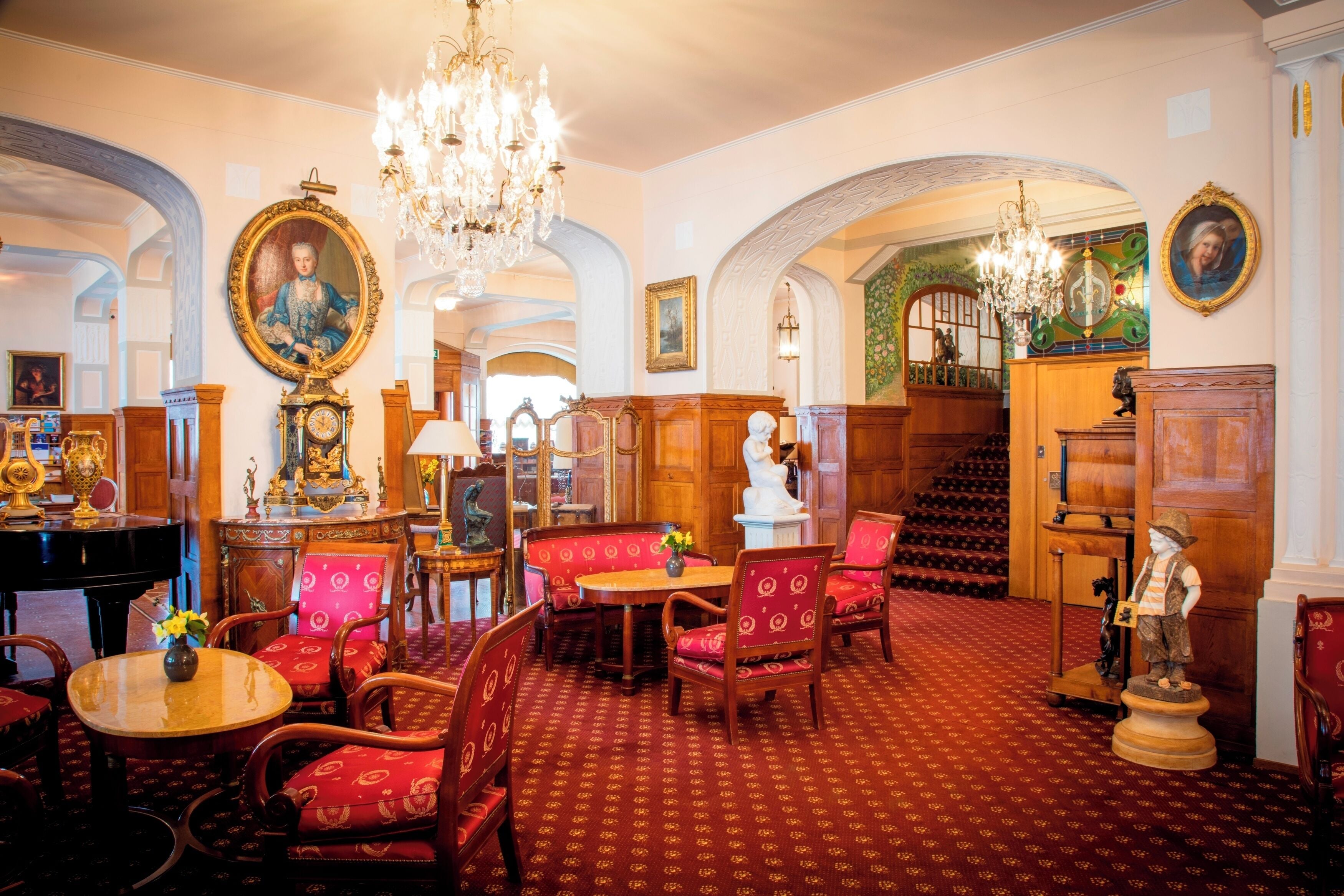 Un'ampia sala con tappeto rosso, arredi in legno, un lampadario e una scala che conduce a un piano superiore.
