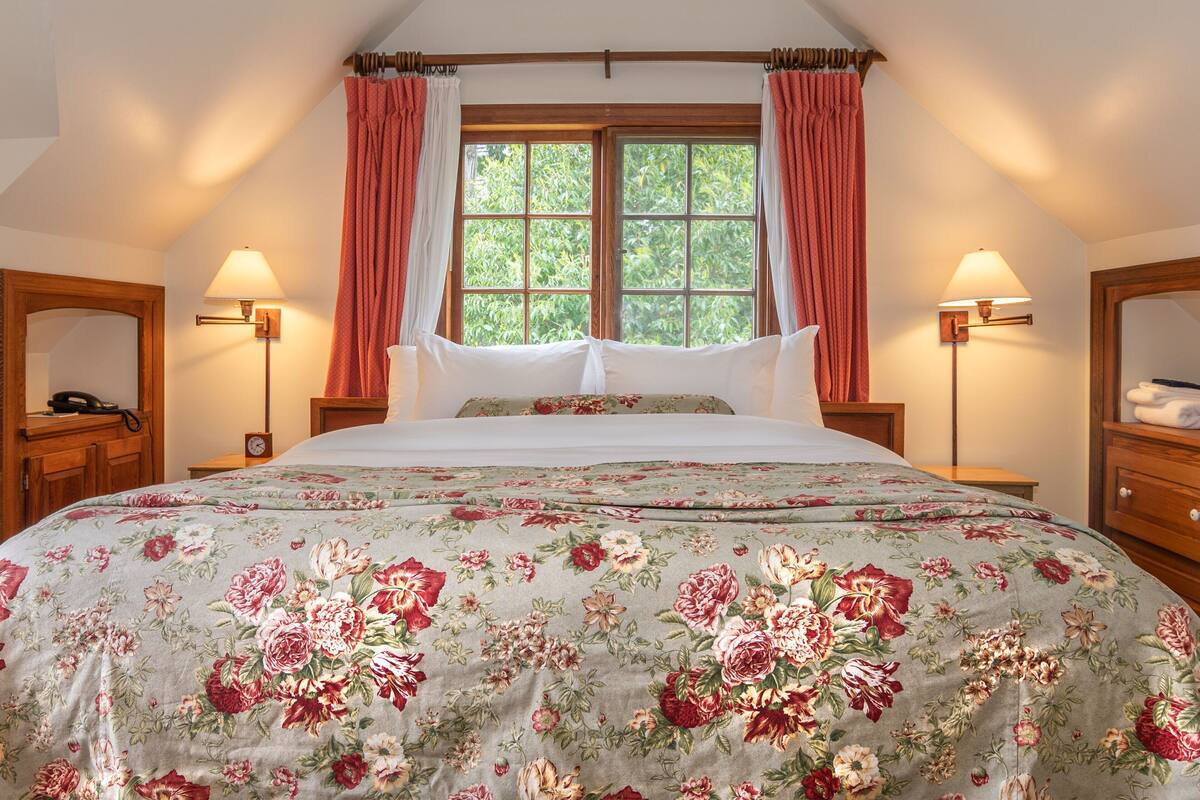 A bedroom with a floral bedspread, two bedside lamps, a wooden nightstand, and a window with curtains.