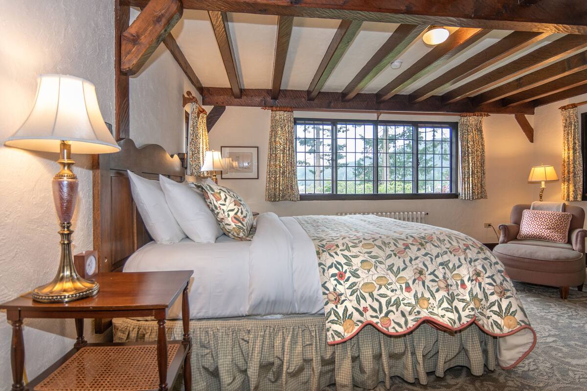 A bedroom with a large bed, two bedside lamps, a wooden headboard, a window with curtains, and a small table with a lamp.