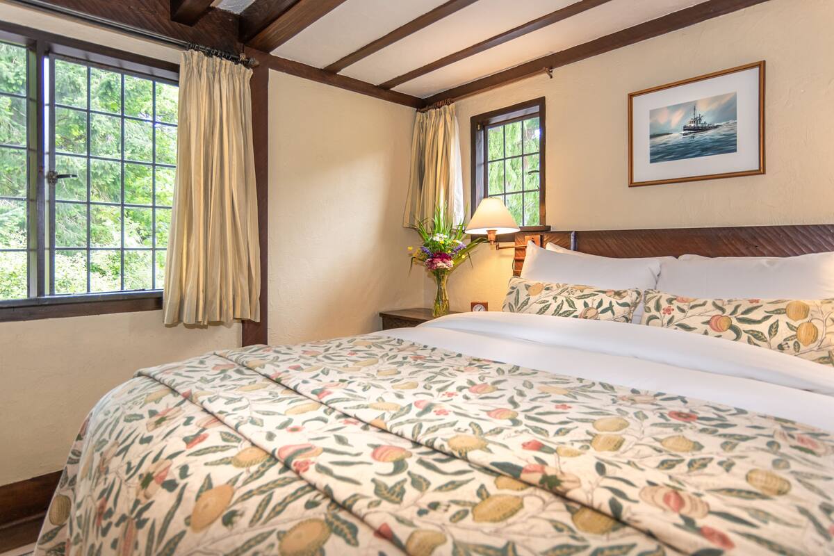 A hotel room with a large bed, floral-patterned bedding, a nightstand with a lamp, a window with curtains, and a framed picture on the wall.