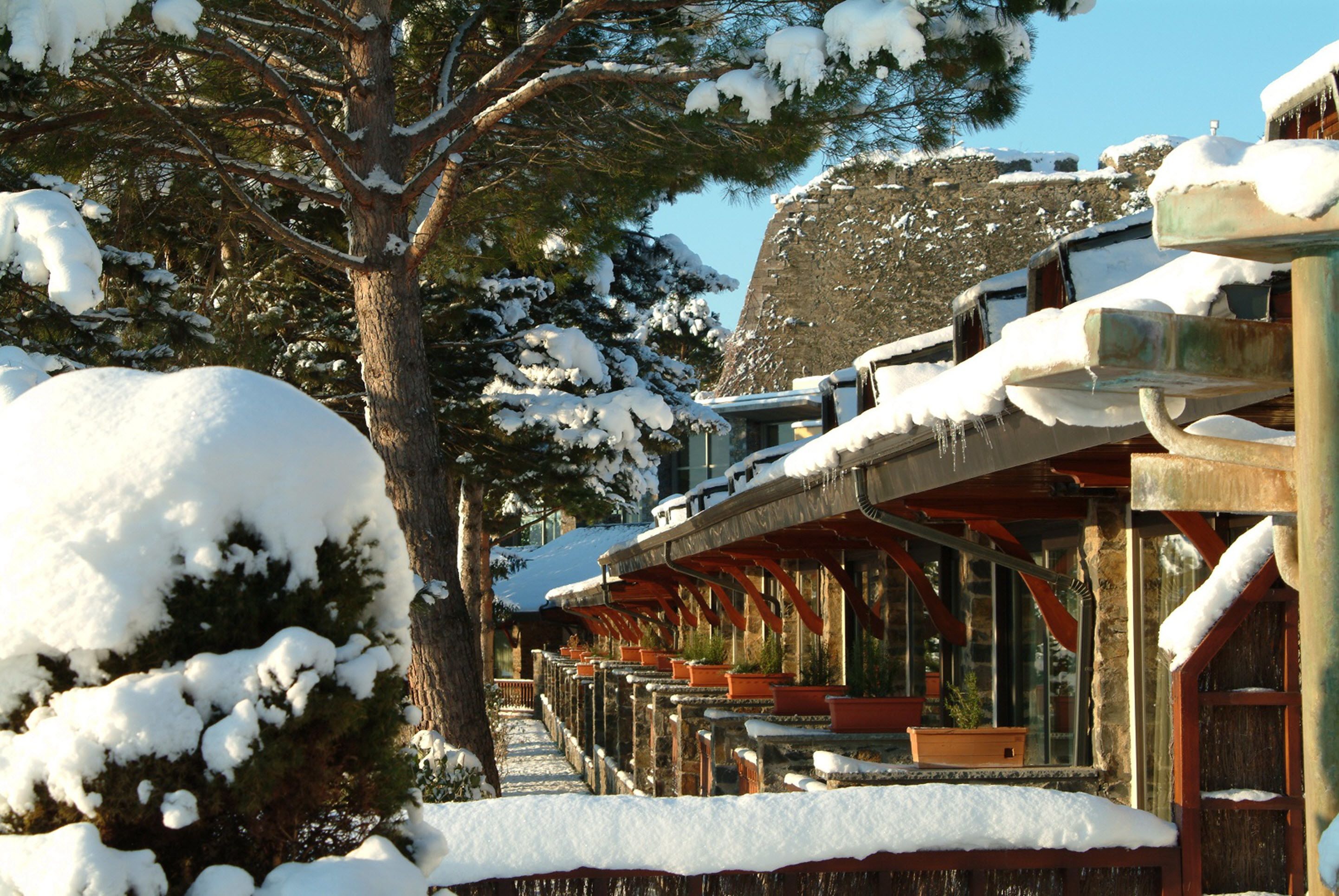A snow-covered outdoor seating area with wooden tables and chairs, surrounded by snow-laden trees and a stone building.