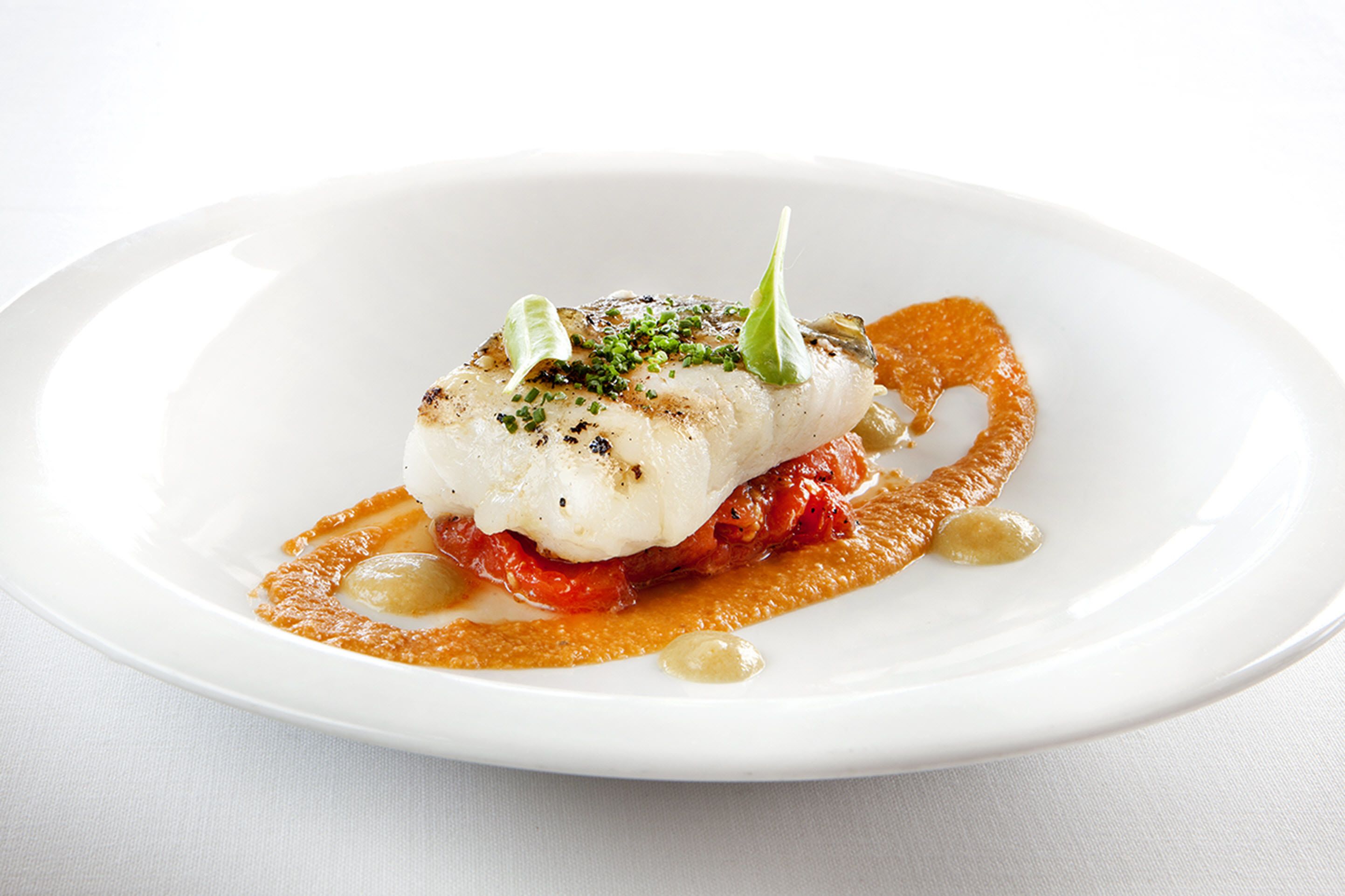A white plate with a piece of fish topped with herbs, accompanied by a sauce and garnishes.