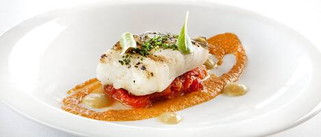 A white plate with a piece of fish topped with herbs, accompanied by a sauce and garnishes.