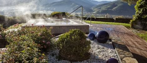 Un jacuzzi rodeado de un jardín con guijarros blancos, una terraza de madera y un telón de fondo montañoso.