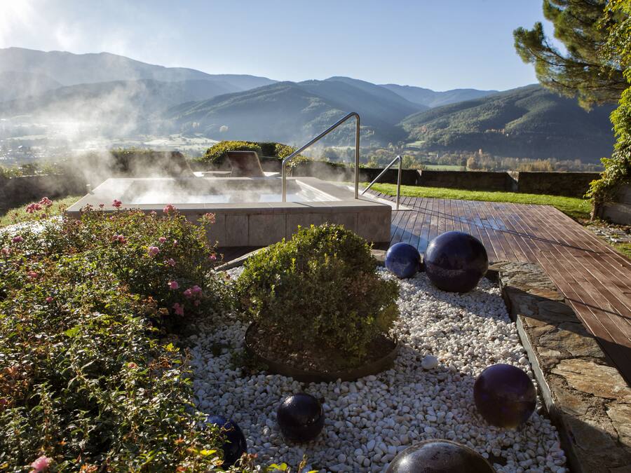 Un jacuzzi rodeado de un jardín con guijarros blancos, una terraza de madera y un telón de fondo montañoso.