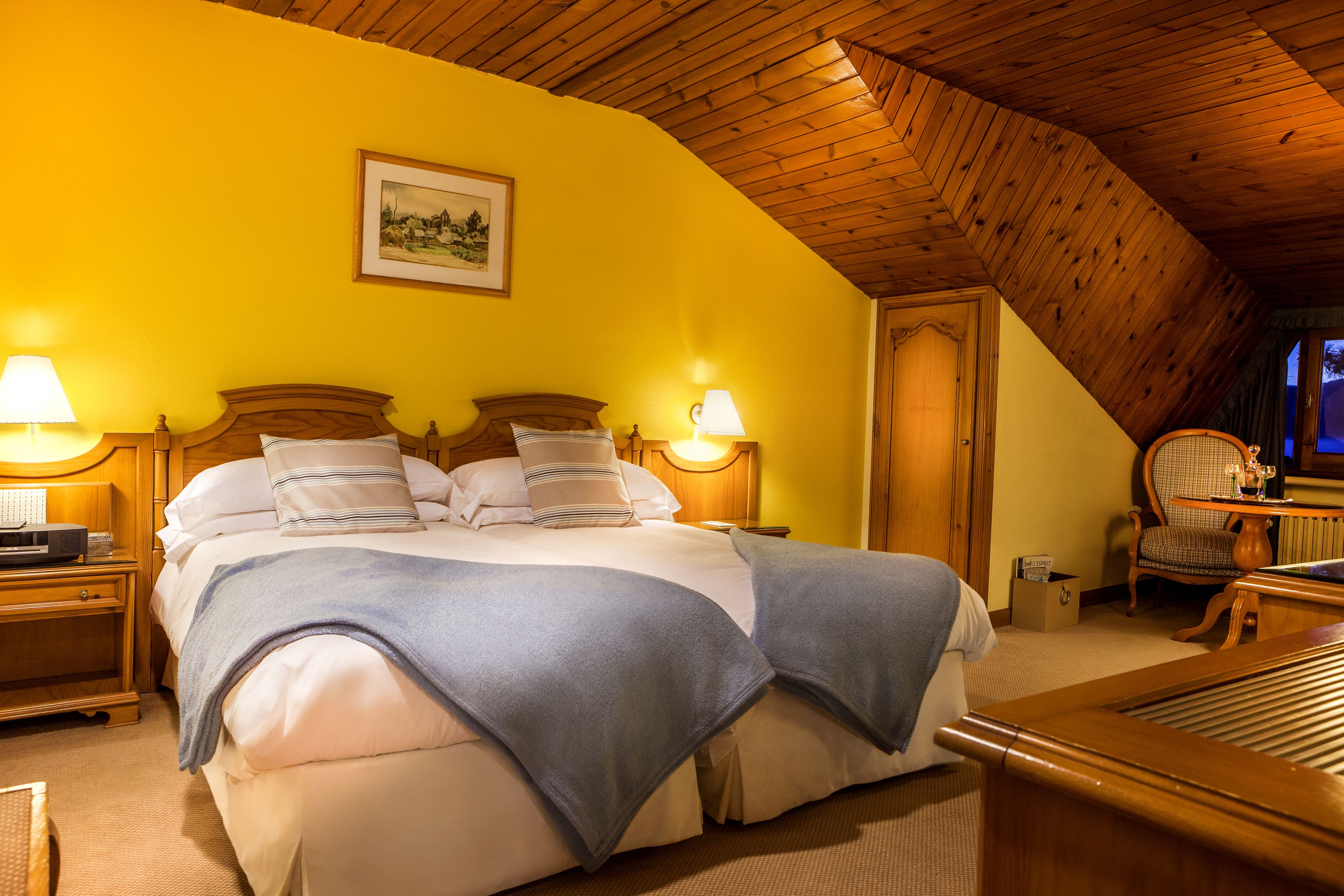 Un dormitorio acogedor con techo de madera, una cama con ropa blanca, dos lámparas de noche y un cuadro en la pared.