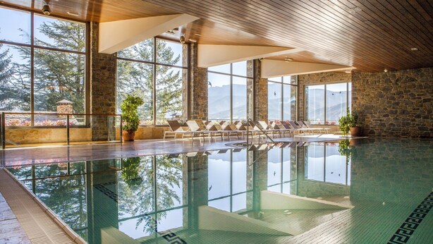 An indoor swimming pool with clear glass walls, stone walls, and wooden ceiling beams.
