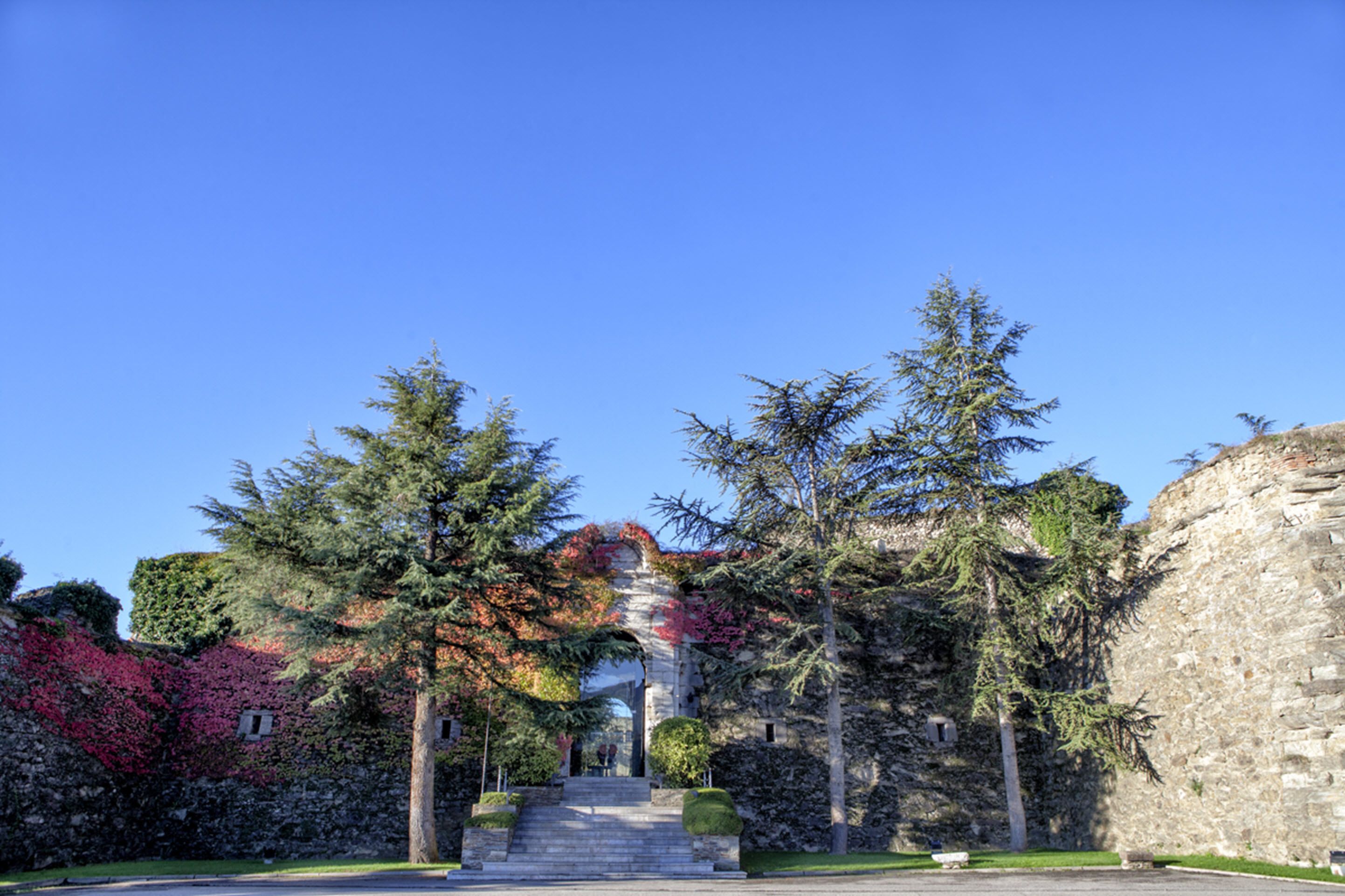 A stone building with a staircase leading to a covered entrance, surrounded by trees and greenery.