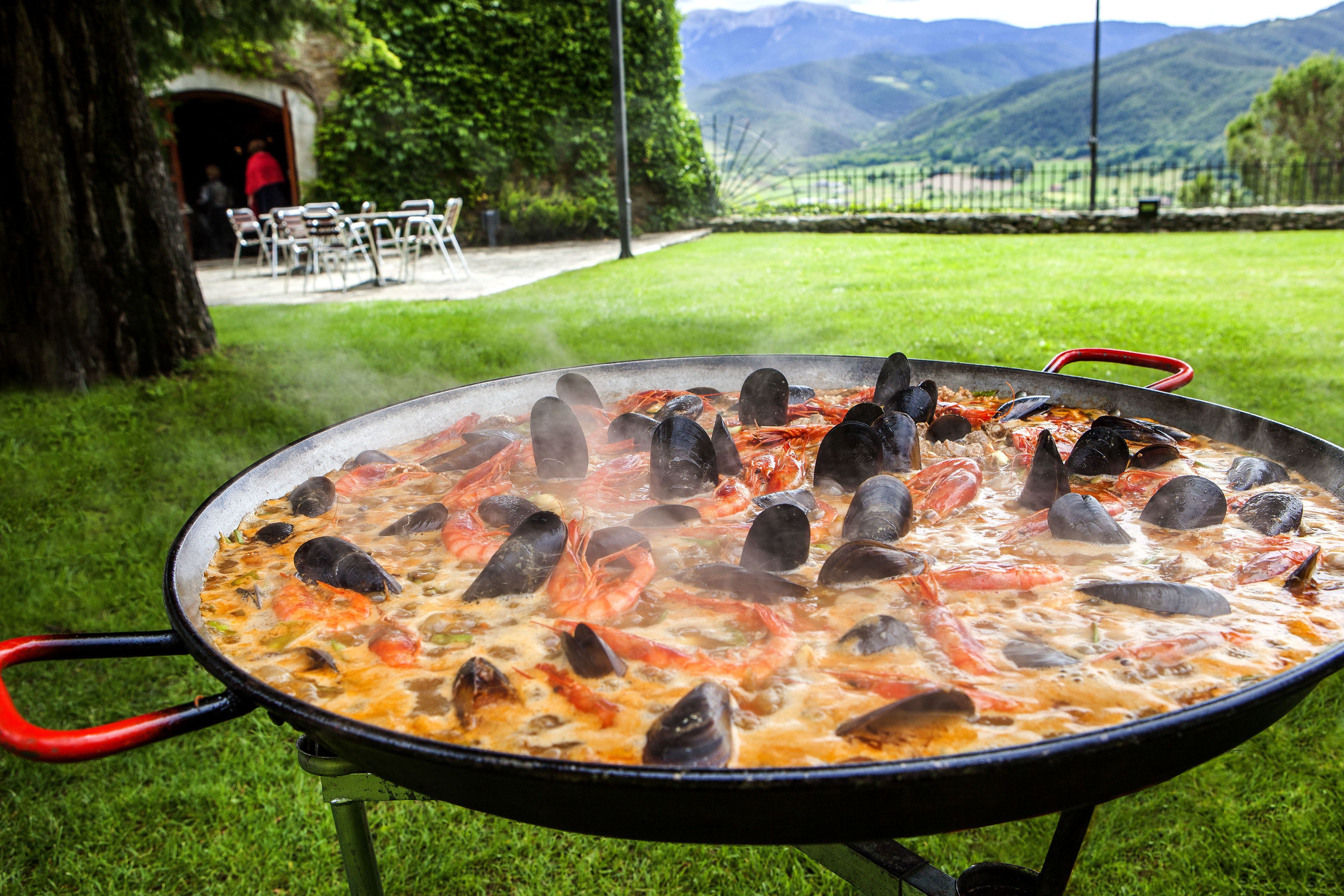 A large outdoor cooking pot filled with seafood, surrounded by a grassy area and a view of mountains.