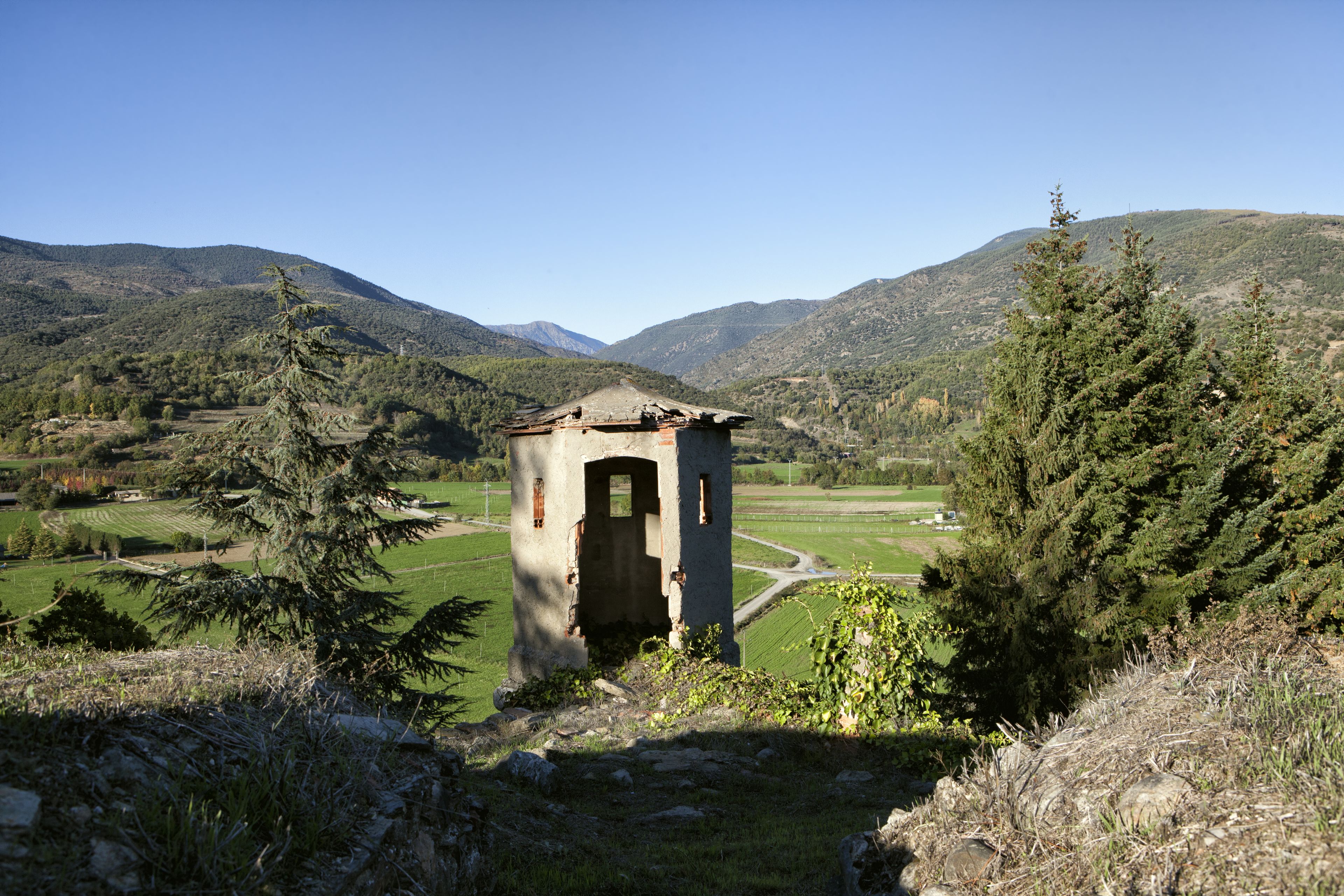 A rustic stone structure with a small door, surrounded by lush greenery and mountains in the background.