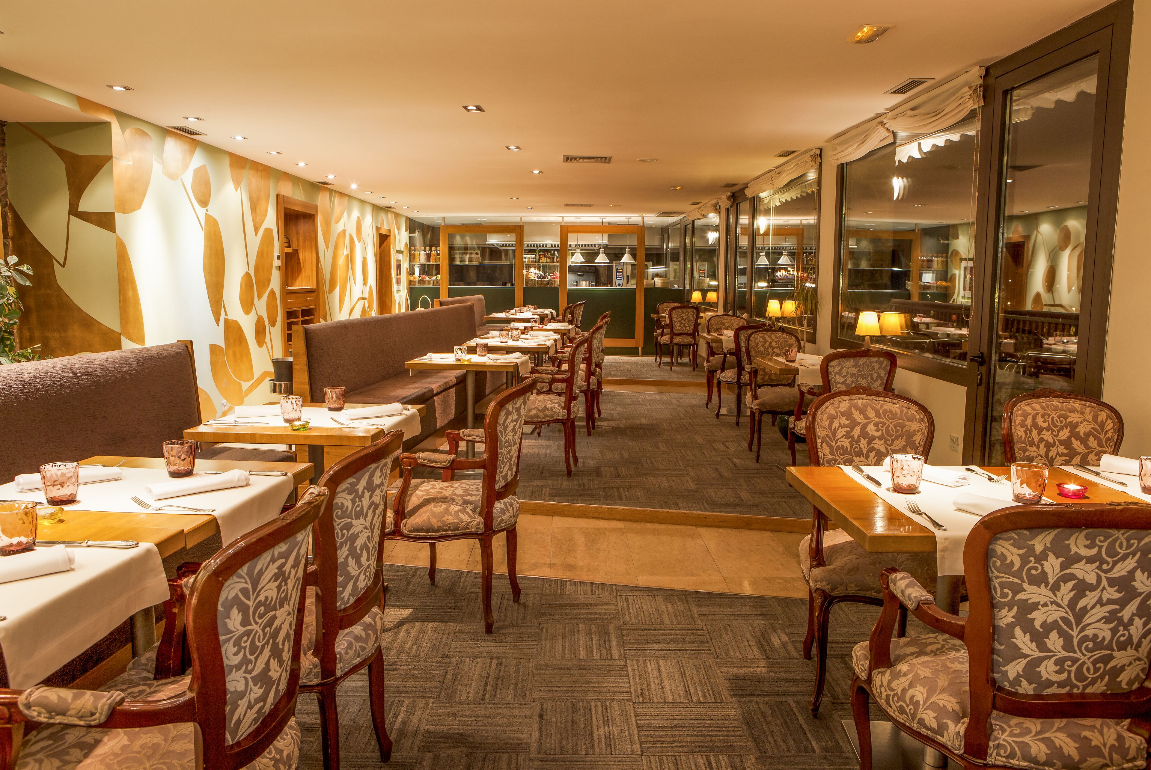 A restaurant with wooden tables and chairs, a patterned wall, and a view of the kitchen through large windows.