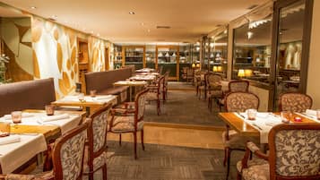 A restaurant with wooden tables and chairs, a patterned wall, and a view of the kitchen through large windows.