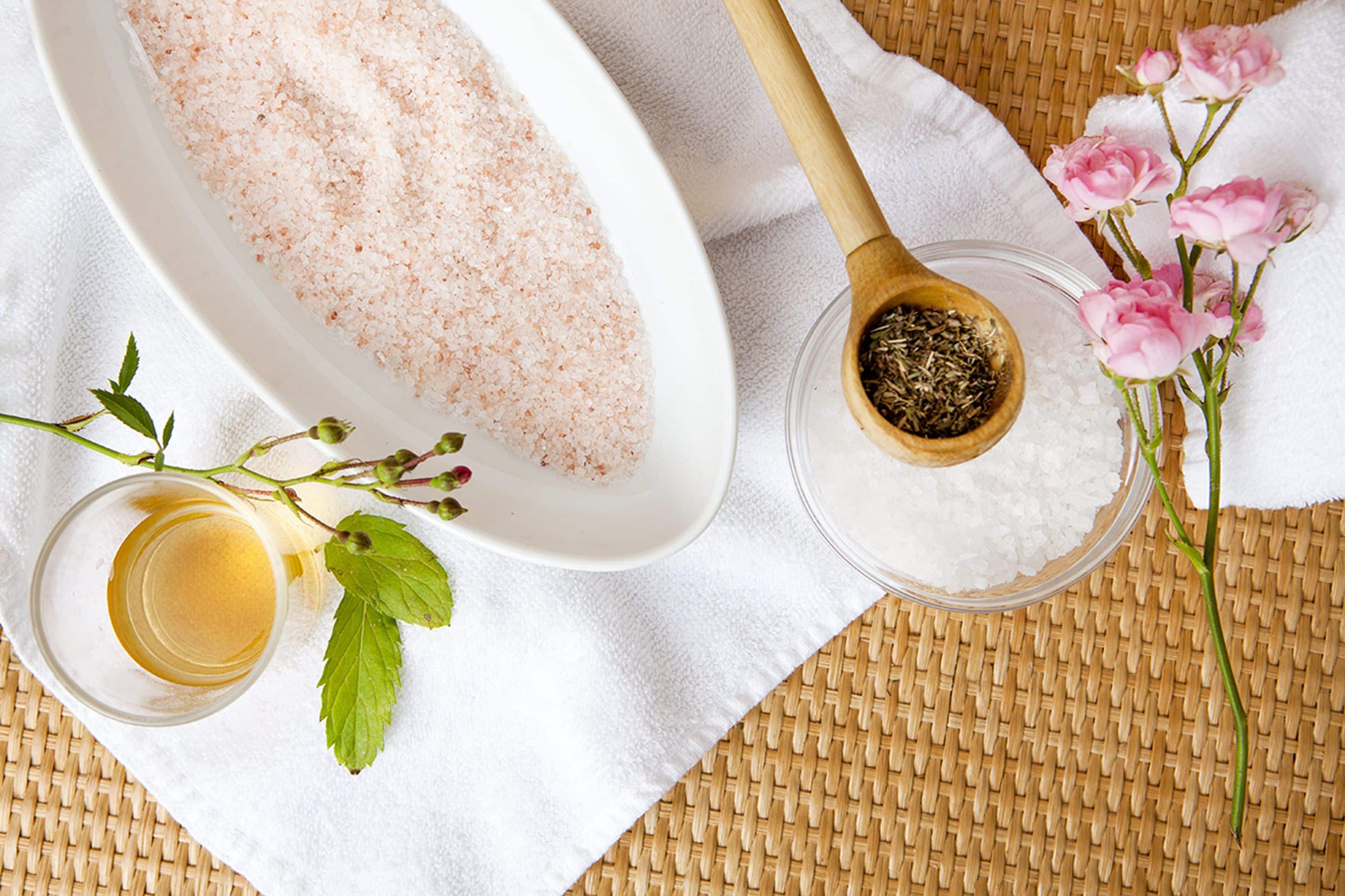 A white bowl filled with pink bath salts, a small glass of liquid, and a wooden spoon with dried herbs.