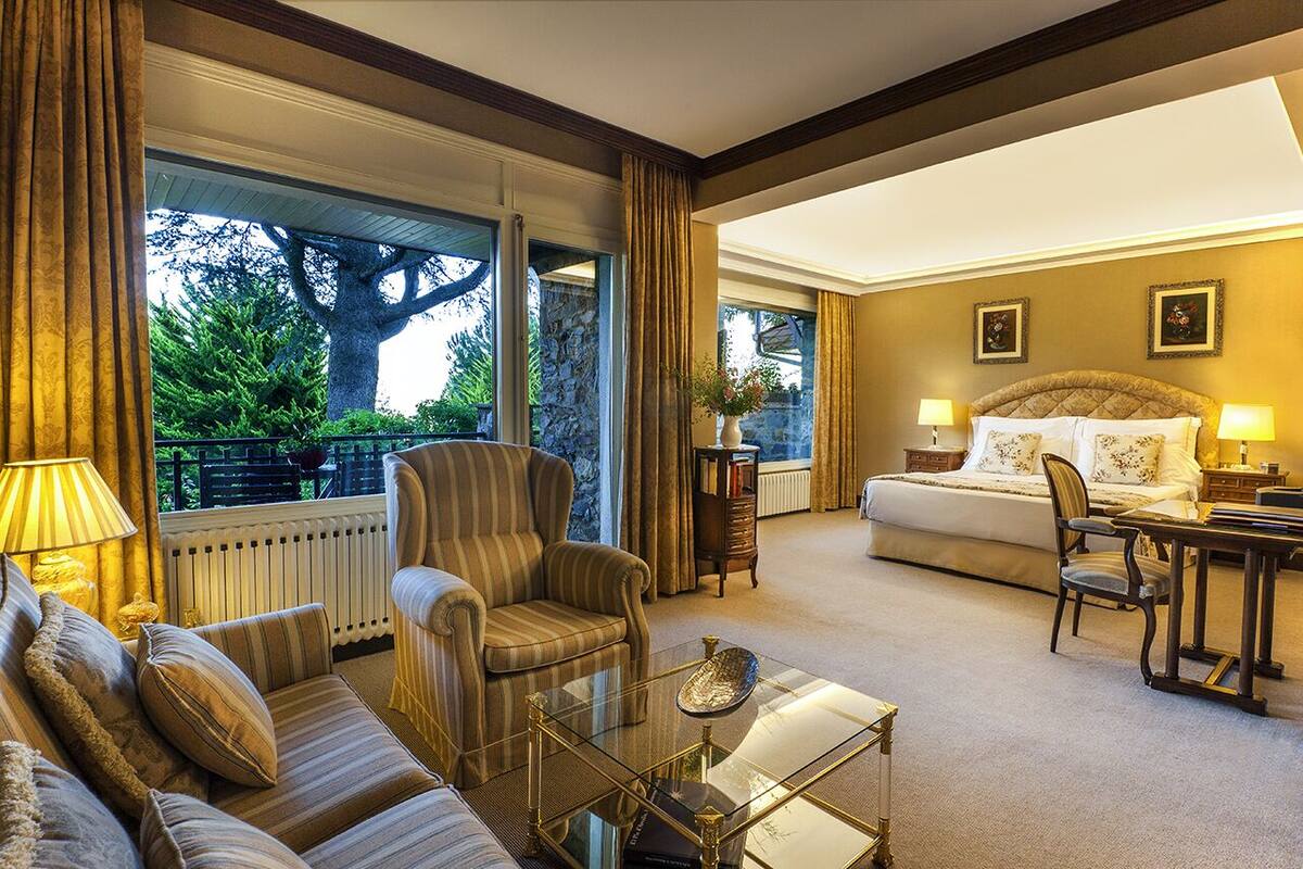 A hotel room with a large bed, a sitting area with a sofa and armchair, a glass coffee table, and a view of the outdoors.