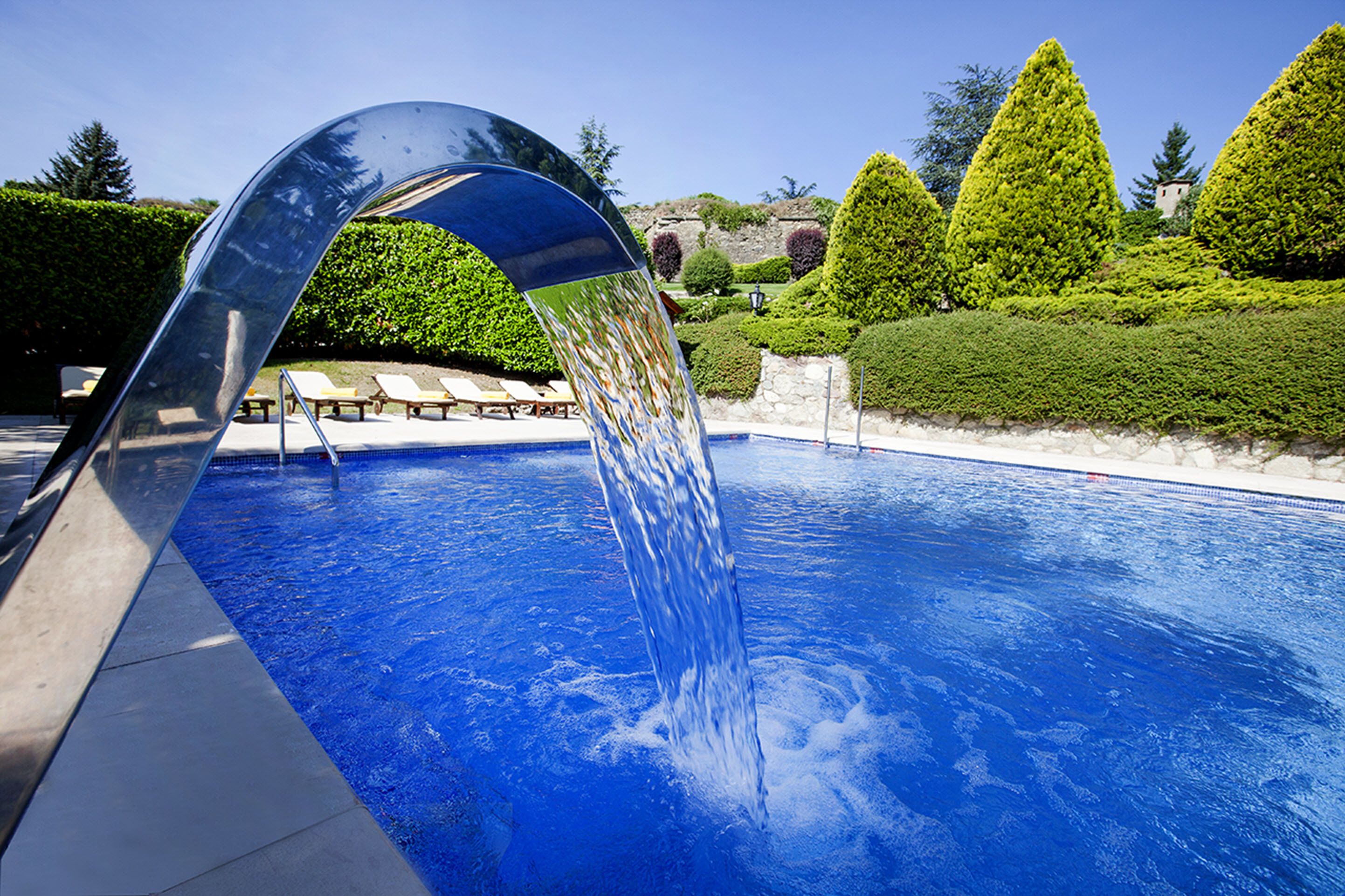 A swimming pool with a modern water feature, surrounded by neatly trimmed hedges and a clear blue sky.