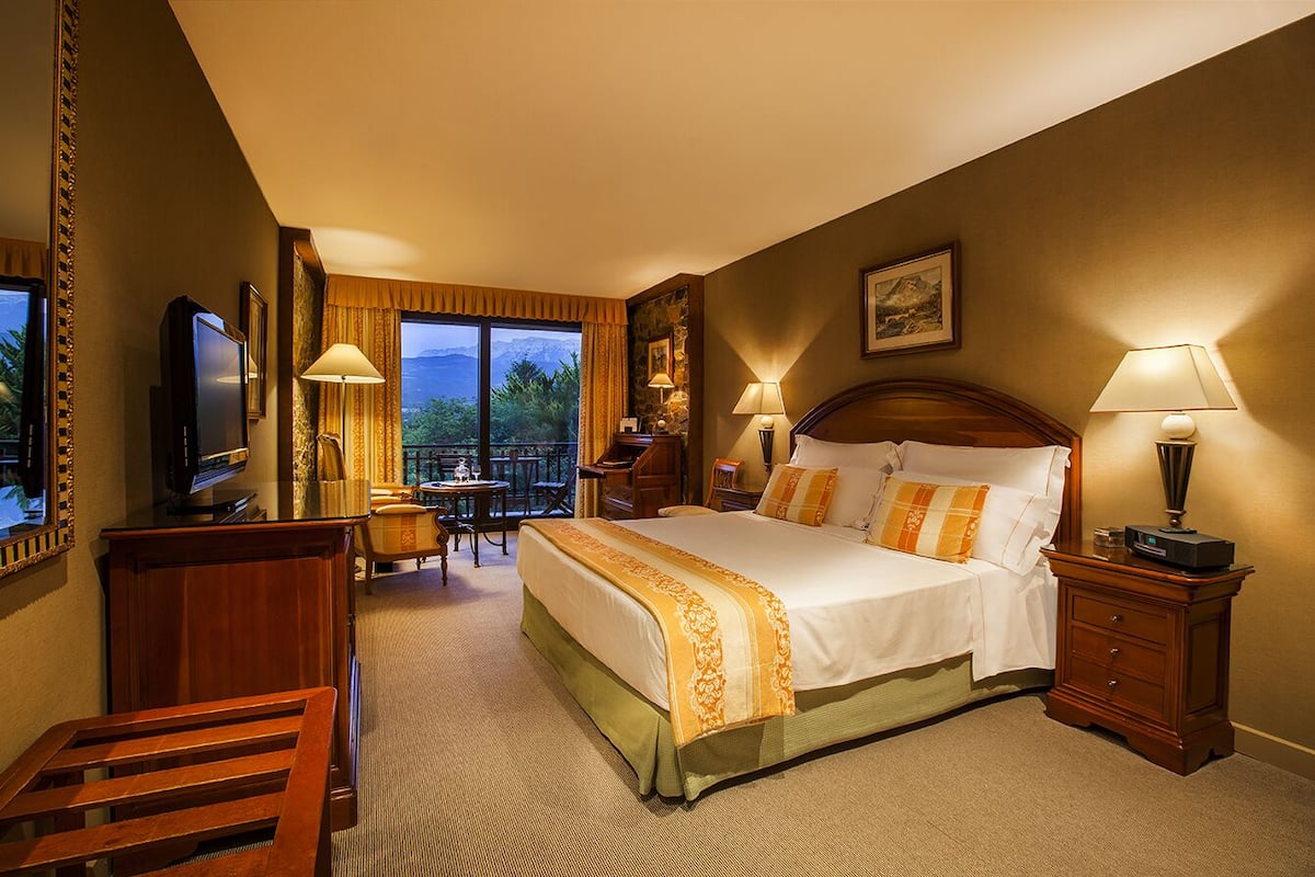 A hotel room with a large bed, a desk, a chair, a TV, and a balcony with a view of the mountains.
