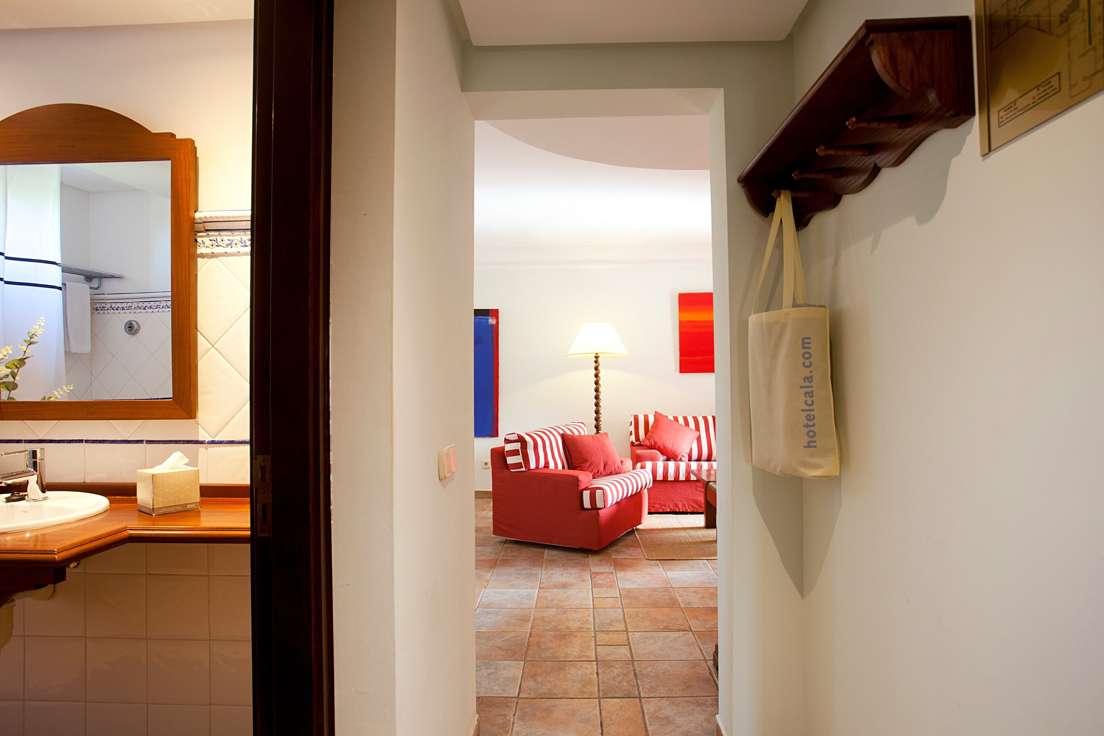A hotel room with a bathroom, a red sofa, a lamp, and a wall-mounted bag.