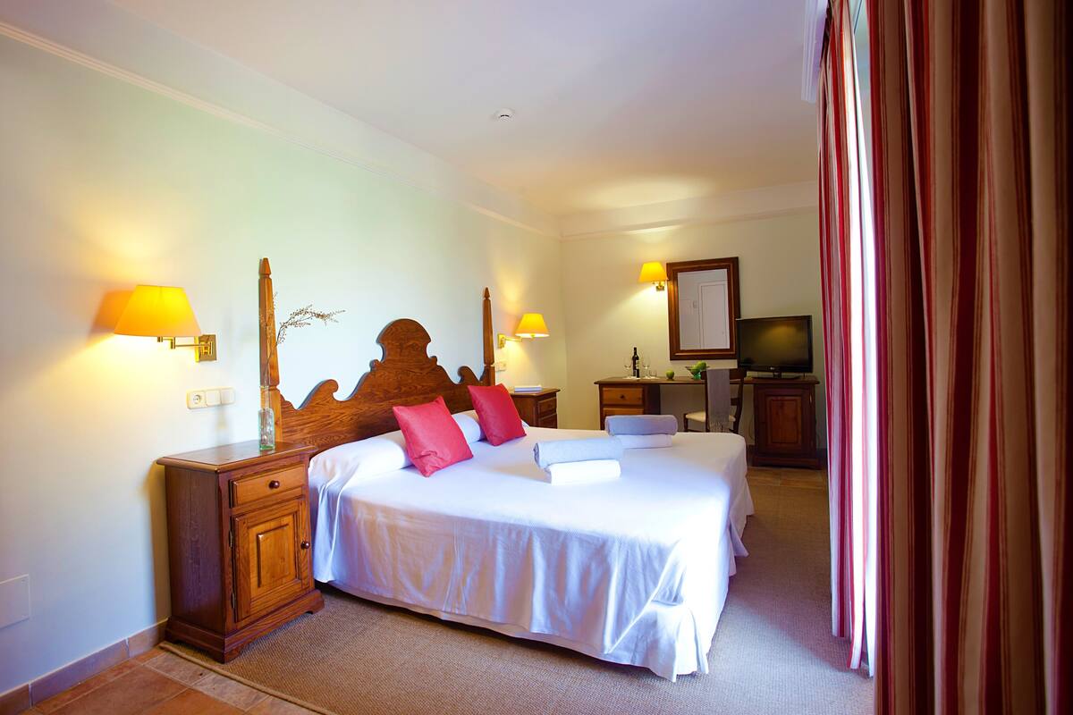 A hotel room with a large bed, bedside lamps, a wooden headboard, a desk with a chair, a television, and a mirror.