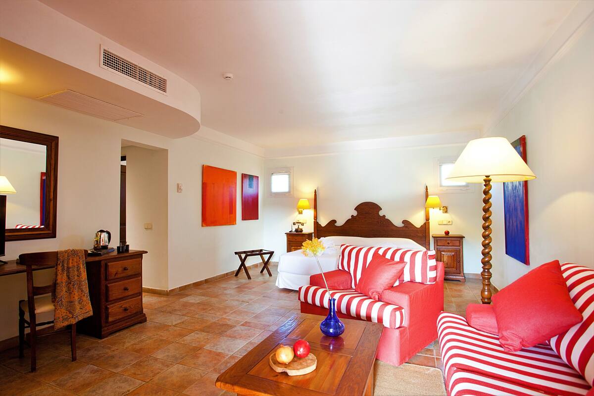 A hotel room with a bed, a red and white striped sofa, a wooden coffee table with fruits, a desk with a chair, and a dresser.