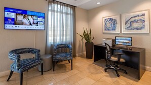 Un espace d’accueil d’hôtel doté d’un téléviseur à écran plat affichant des informations sur l’hôtel, de deux fauteuils à motifs, d’un bureau avec un ordinateur et de deux tableaux abstraits encadrés accrochés au mur.