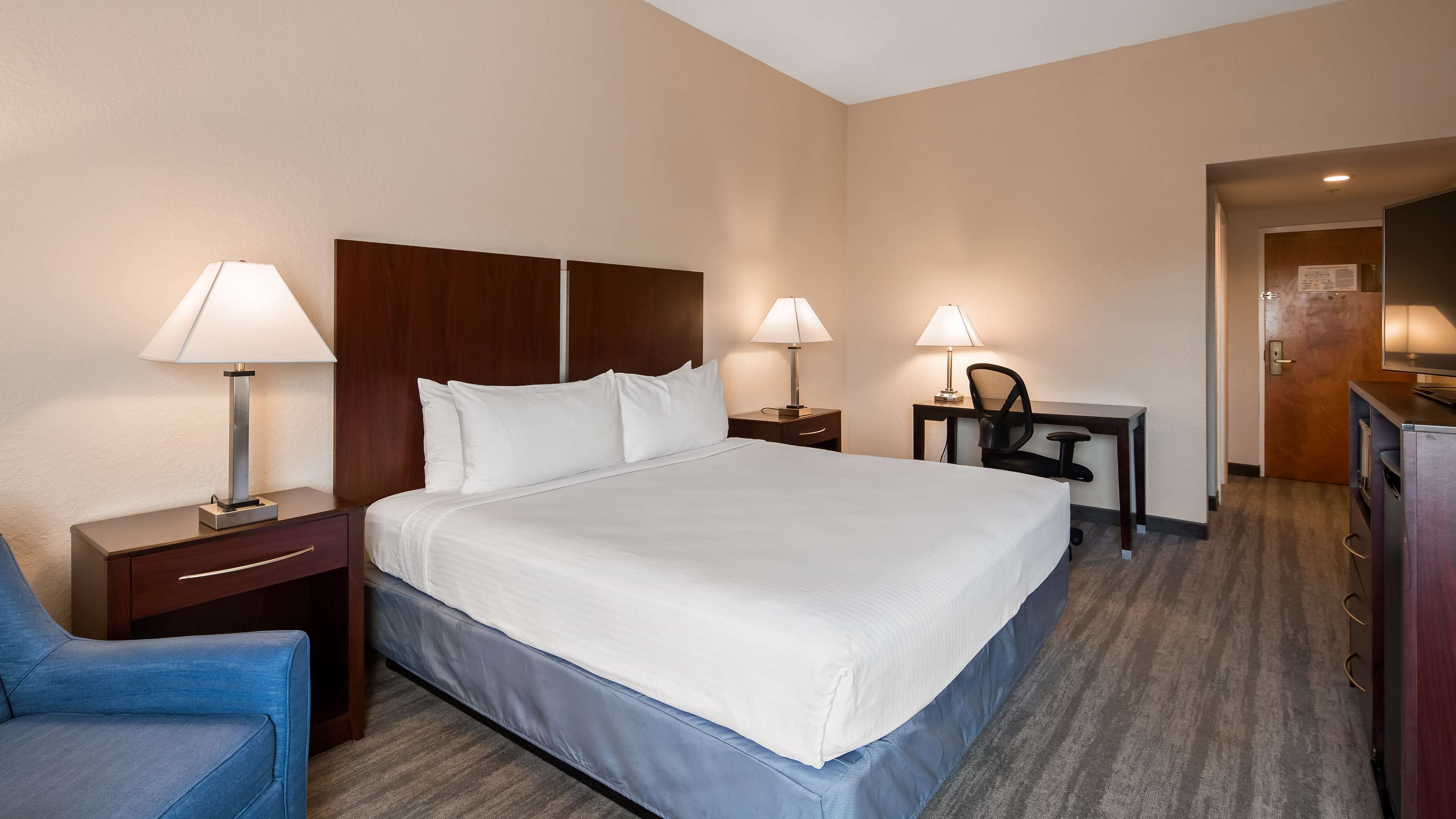 A hotel room with a large bed, two bedside tables with lamps, a desk with a chair, and a blue armchair.