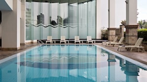 A swimming pool with clear blue water, surrounded by lounge chairs and a glass wall reflecting the sky.