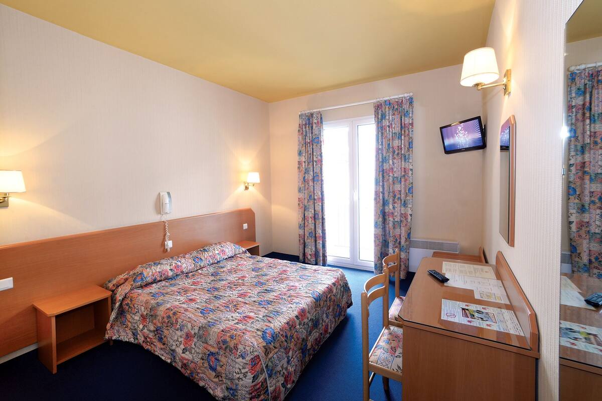 Een hotelkamer met een bed, nachtkastje, lamp, bureau, stoel en een televisie aan de muur.