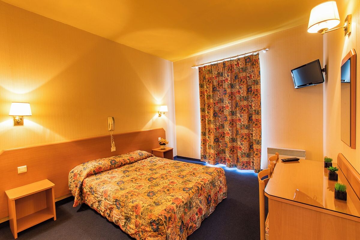 A hotel room with a bed, bedside tables, a desk with a chair, a TV, and a curtain.