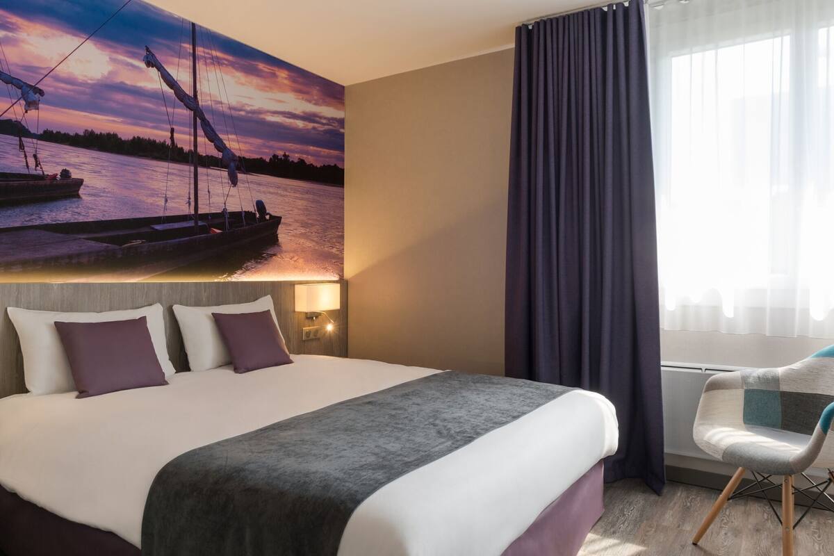 Quarto de hotel com uma cama grande, uma cadeira, uma mesinha de cabeceira e um mural na parede retratando um pôr do sol sobre a água.