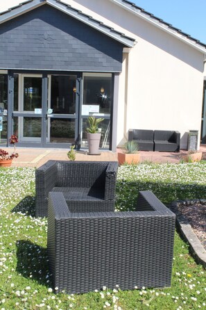 A modern building with a dark roof, large glass doors, and a patio area with wicker furniture and potted plants. - The Originals City, Hôtel La Terrasse, Tours Nord (Tours)