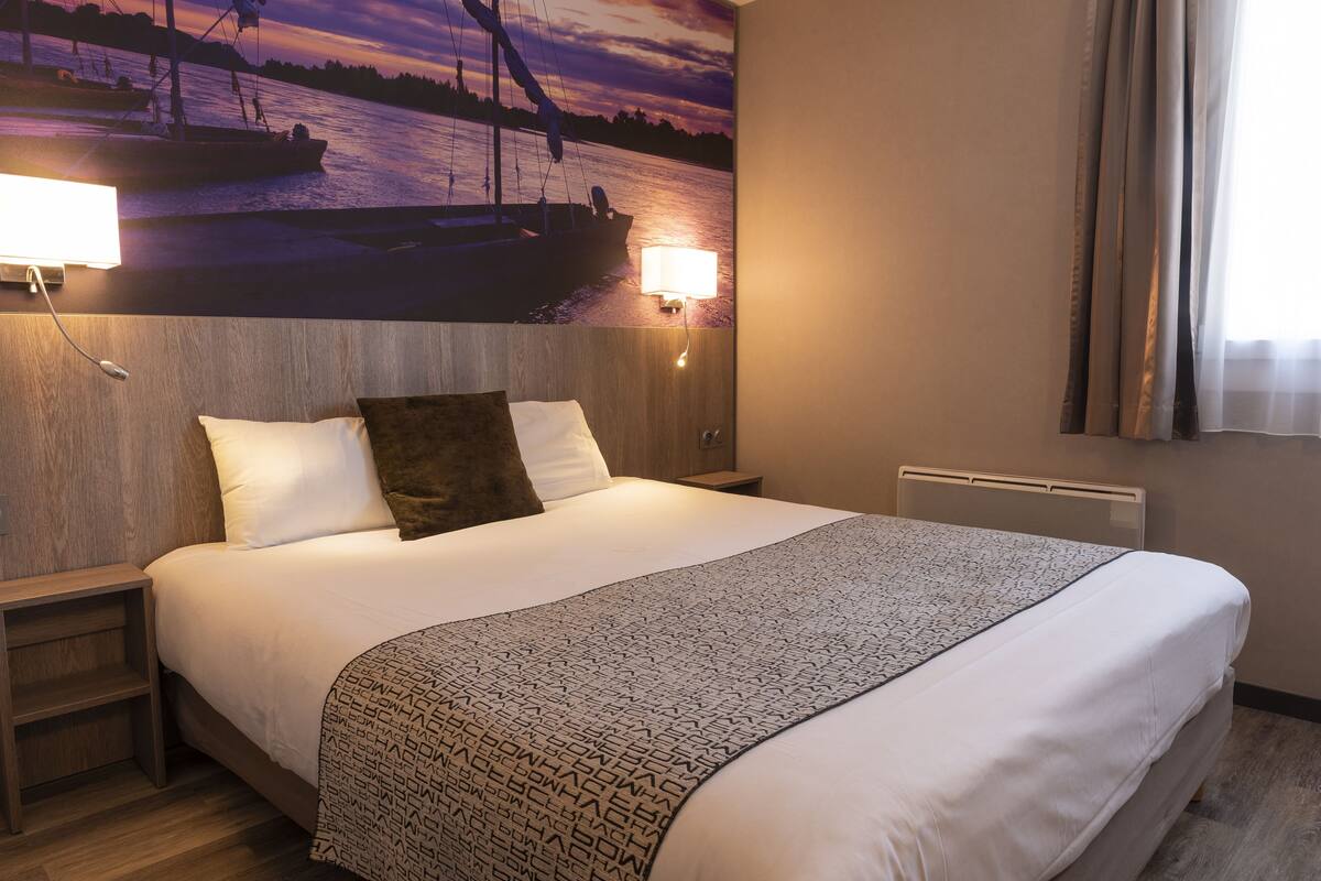 Quarto de hotel com uma cama grande, duas luminárias de cabeceira, cabeceira com um mural de barcos e uma janela com cortinas.