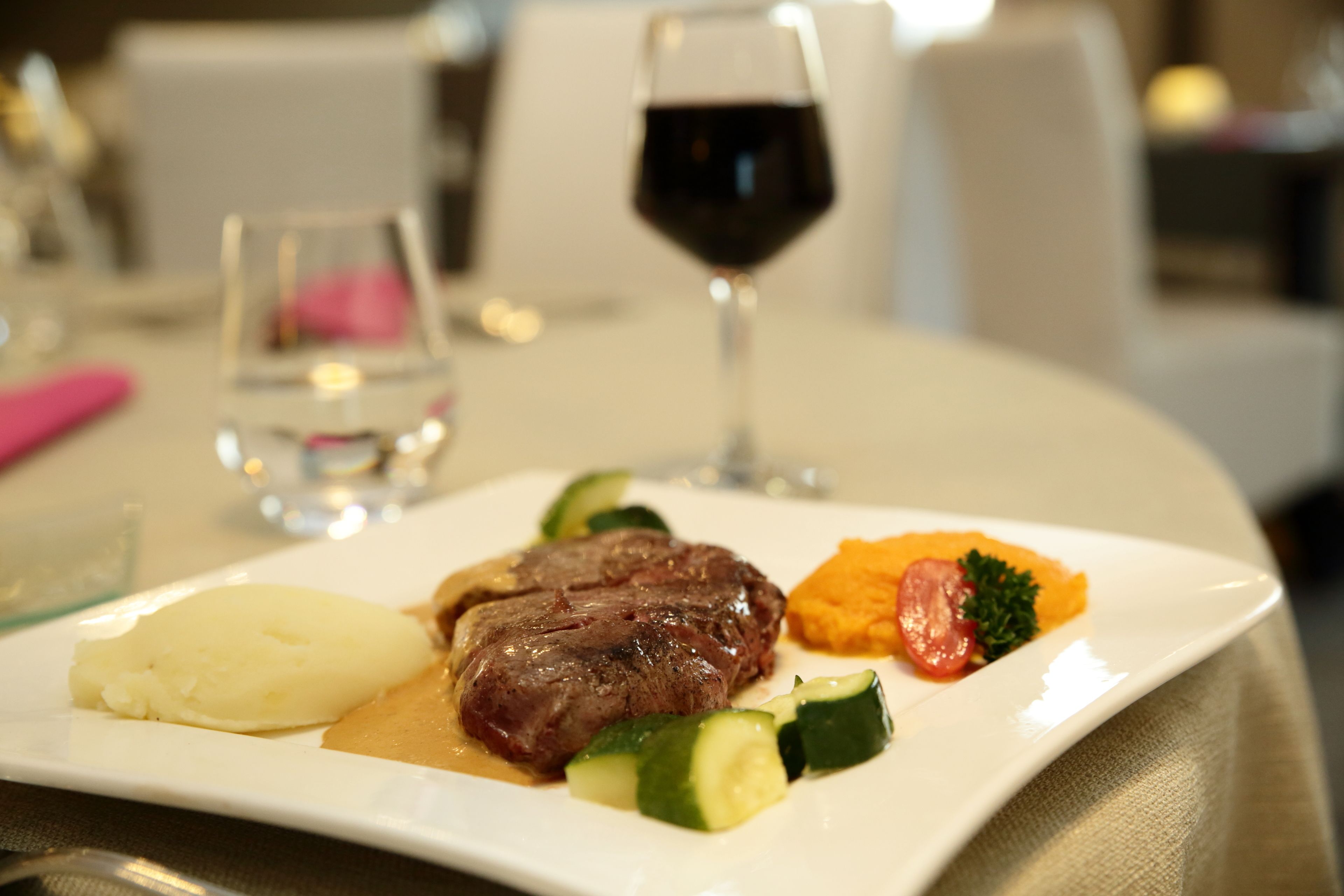 Un plato con filete, puré de papas y verduras, junto a una copa de vino tinto sobre una mesa.