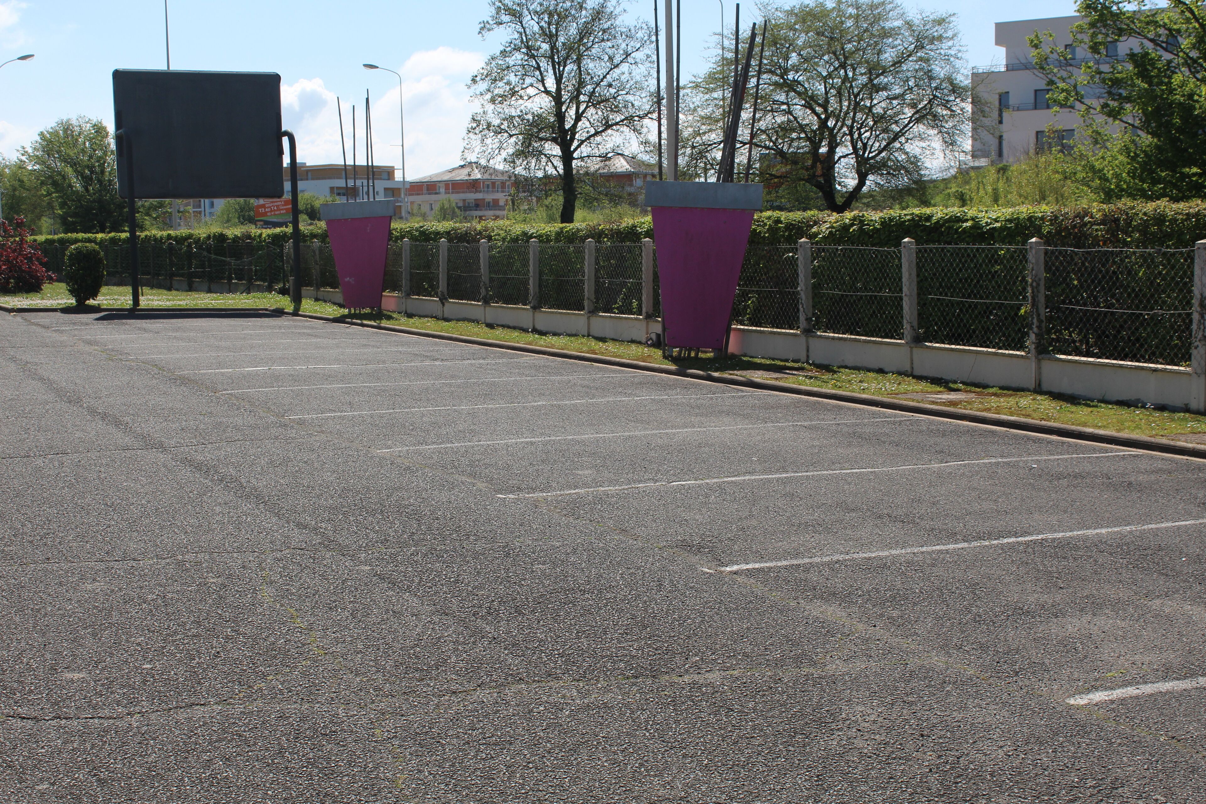 A parking lot with marked spaces, a large blank sign, and a chain-link fence with a hedge.