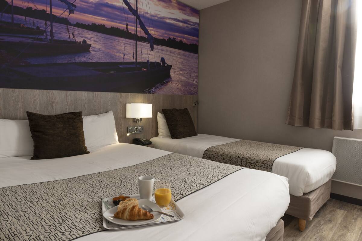 Quarto de hotel com duas camas, um mural na parede com um pôr do sol sobre a água, uma bandeja com croissants e suco, e um abajur na mesa de cabeceira.