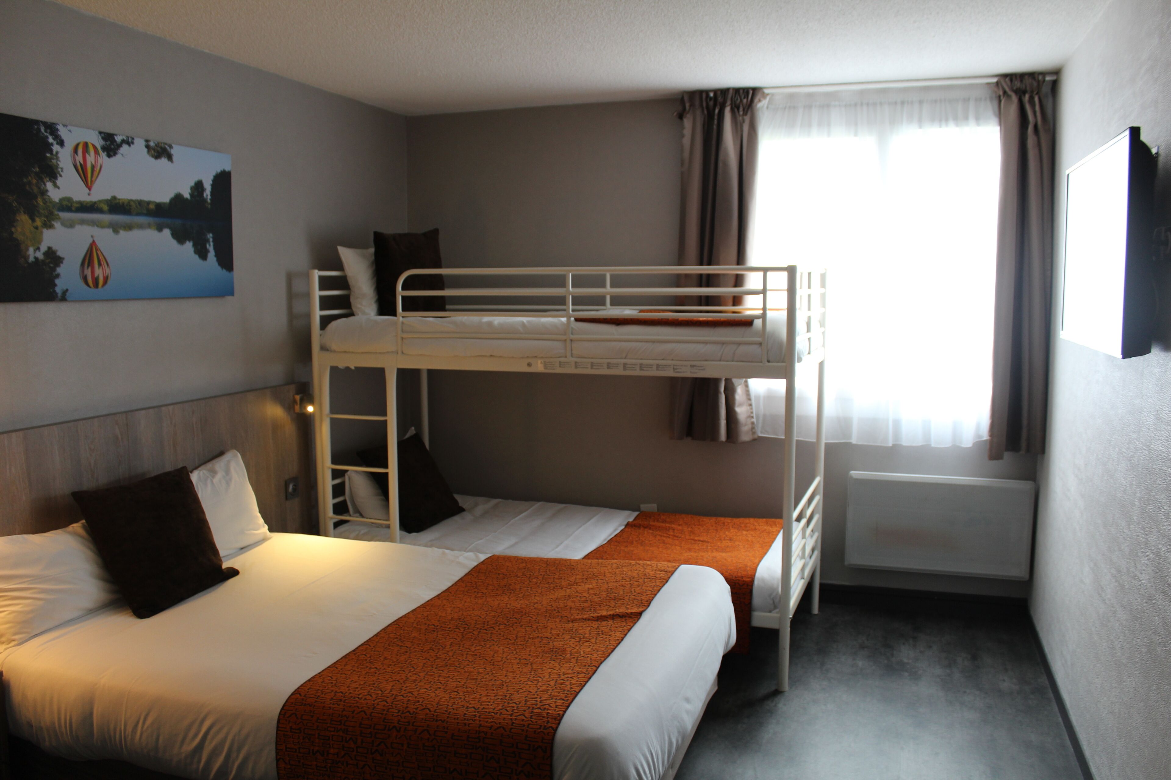 Una habitación de hotel con una cama litera, una cama individual, un cuadro en la pared, una ventana con cortinas y un radiador.