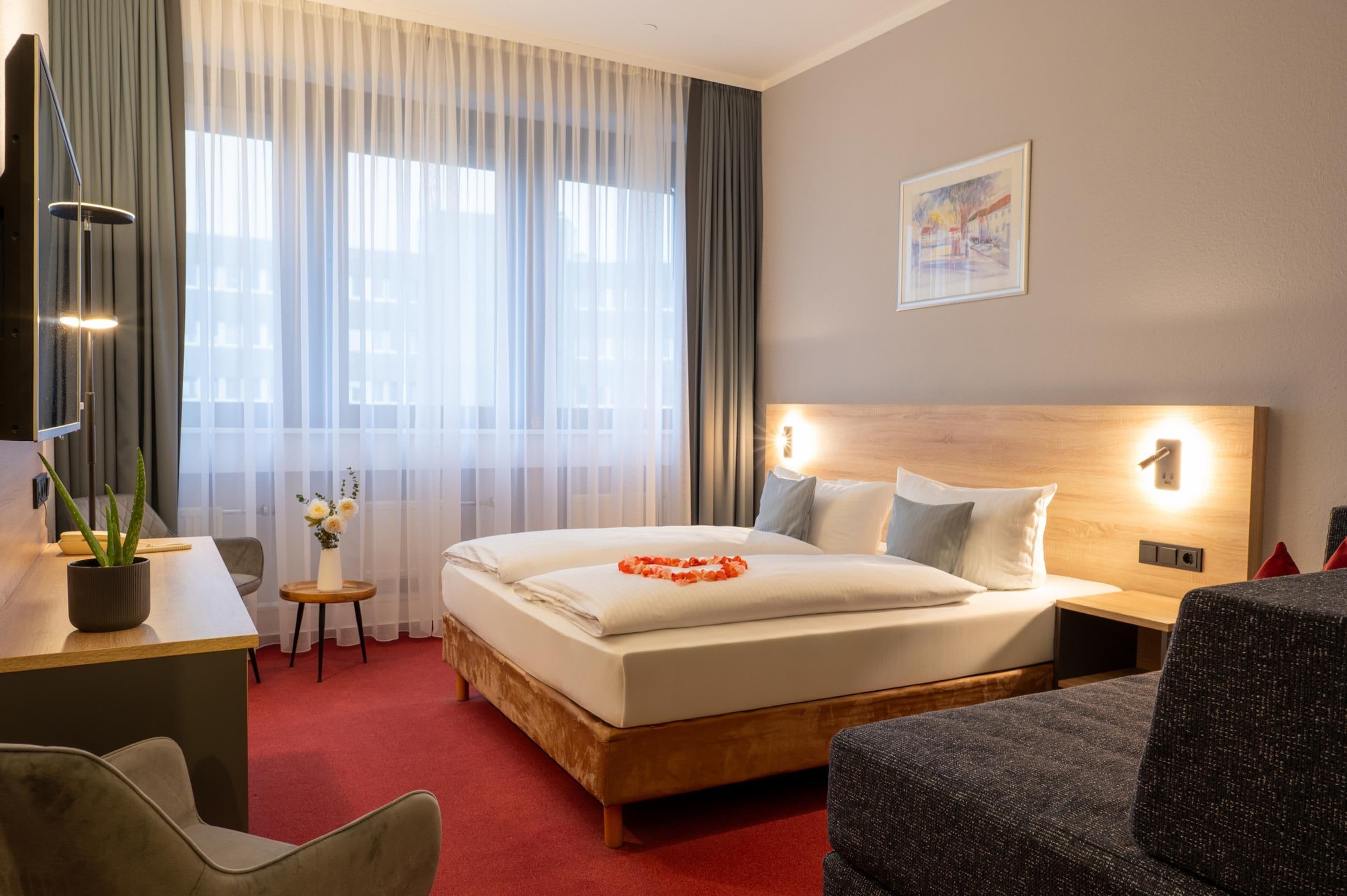 Ein Hotelzimmer mit einem großen Bett, einem Schreibtisch mit Lampe, einem Sessel, einem kleinen Tisch mit Blumen und einem Gemälde an der Wand.