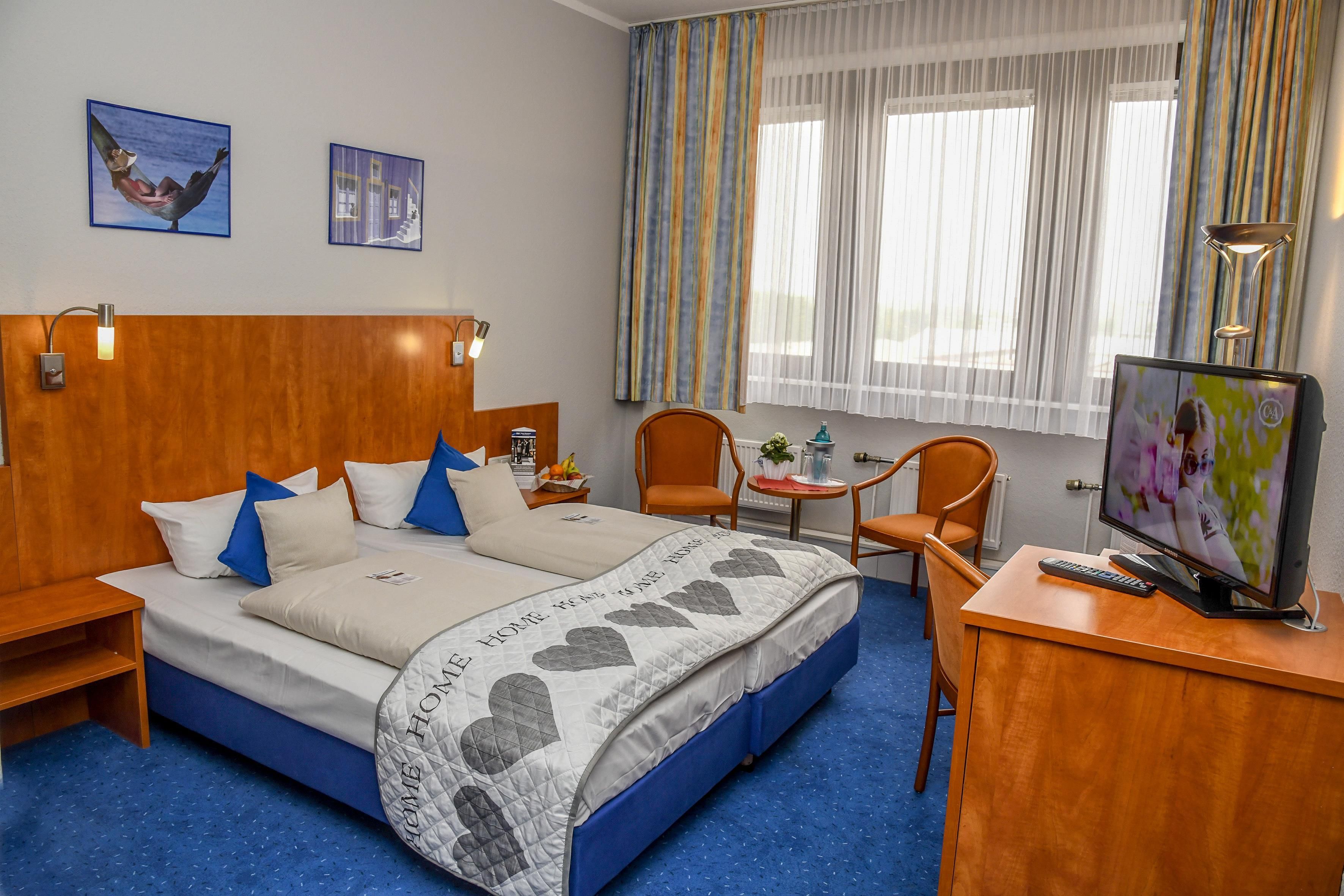 A hotel room with a bed, two chairs, a desk with a TV, and a window with curtains.