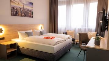 Ein Hotelzimmer mit Bett, Nachttischen, einem Schreibtisch, einem Sessel sowie einem Stadtbild an der Wand.