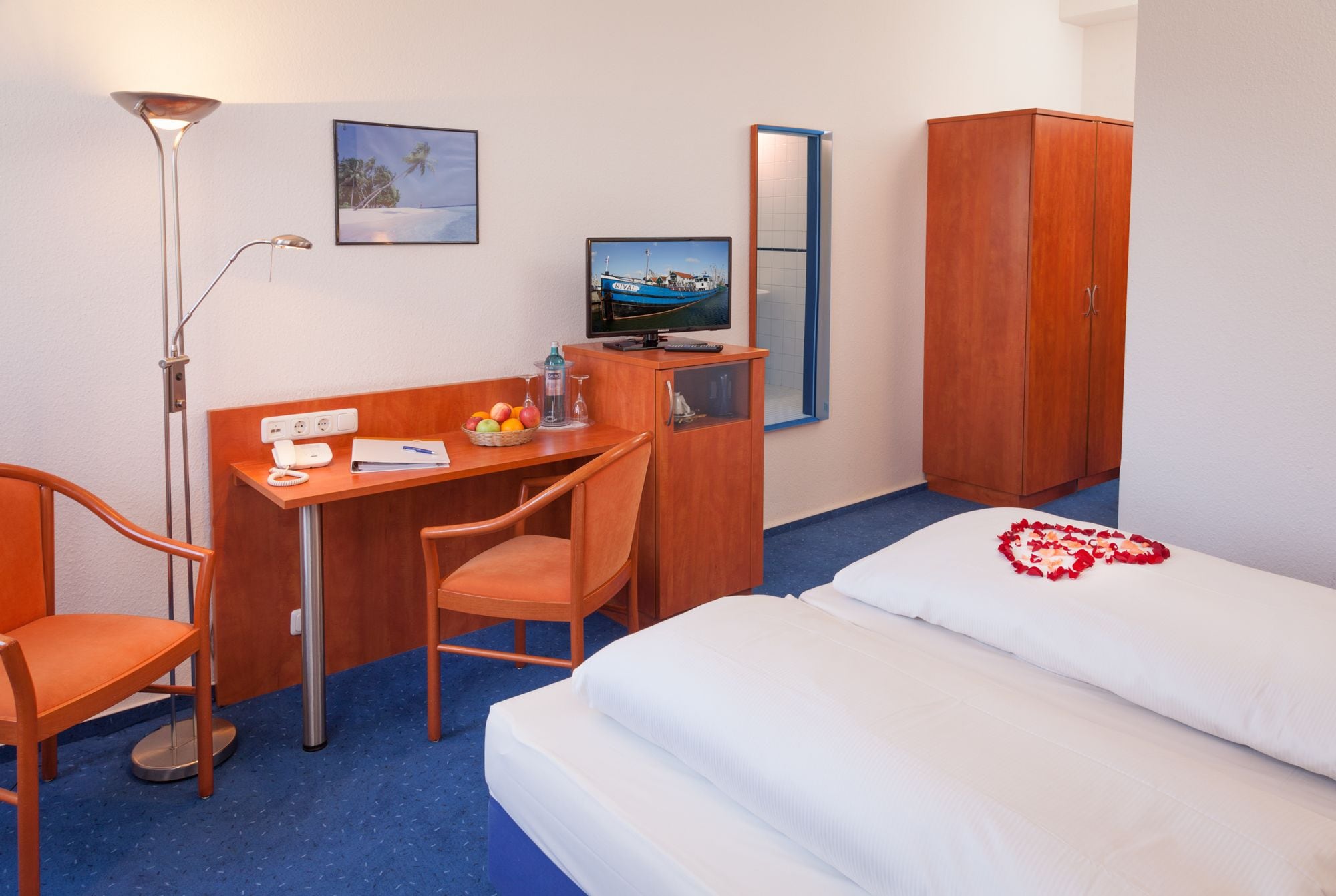Ein Hotelzimmer mit Bett, Schreibtisch mit Stuhl, Kleiderschrank, Schreibtischlampe, Fernseher und einem kleinen Tisch mit Obstkorb und einer Flasche.