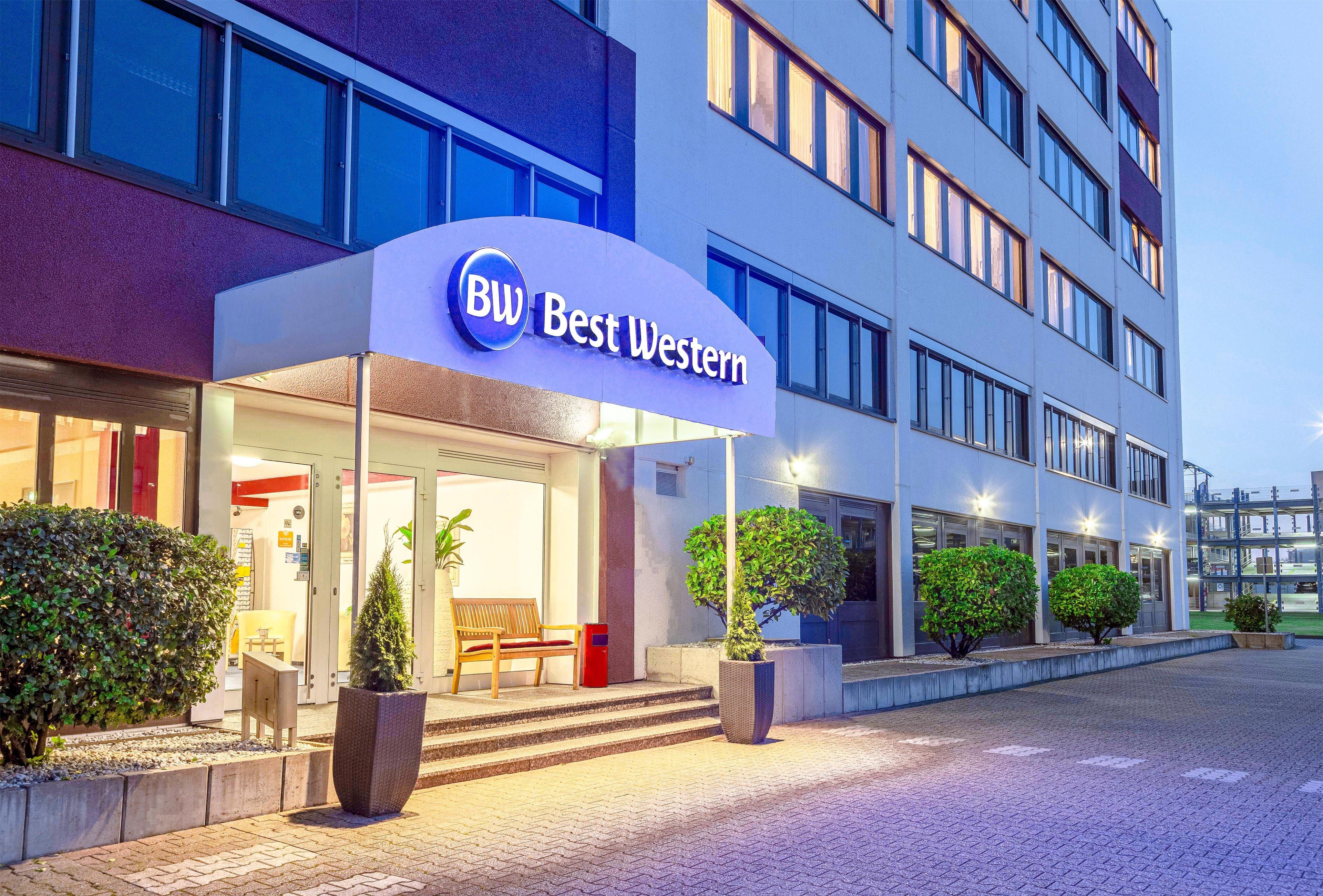 L’entrée d’un hôtel, avec une enseigne indiquant « BW Best Western », un espace de repos abrité avec un banc, et des plantes en pot.
