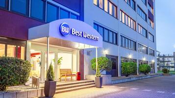 L’entrée d’un hôtel, avec une enseigne indiquant « BW Best Western », un espace de repos abrité avec un banc, et des plantes en pot.
