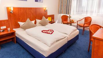 Habitación de hotel con una cama grande, cabecera de madera, mesita de noche con lámpara, pequeña mesa con frutero y dos sillones naranjas.
