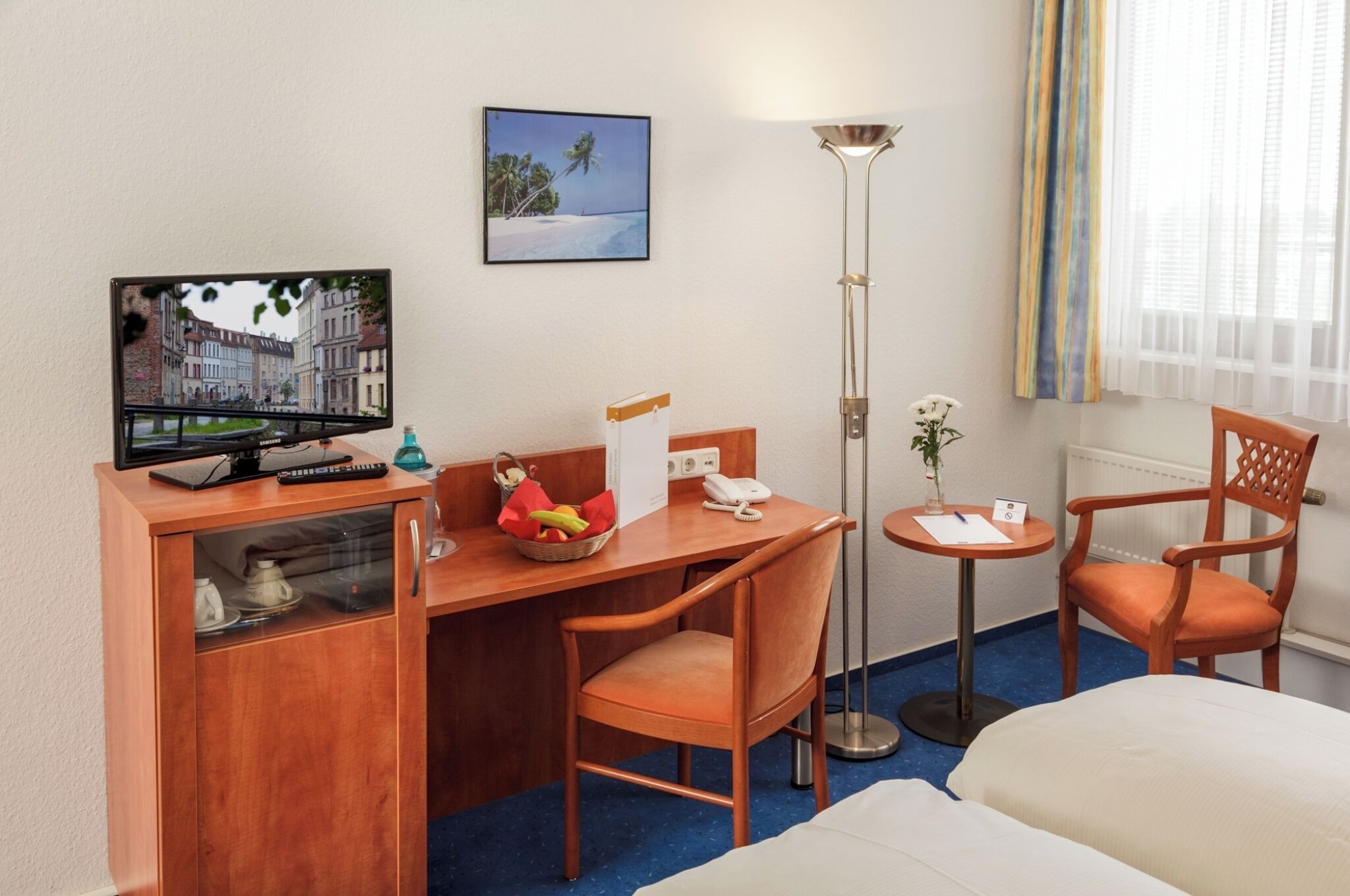 Ein Hotelzimmer mit einem Holztisch, einem Fernseher, einem Bett, einem Sessel, einem kleinen Tisch mit einer Lampe und einem Bild an der Wand.